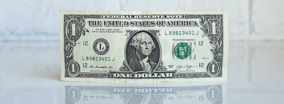 A green dollar bill with George Washington's face sits upright against a white brick wall reflects on a clear counter