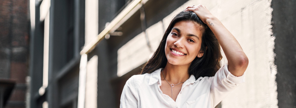 Brunette woman wearing a white shirt smiles with dental veneers and her hand on her head in front of an old building