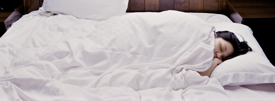 Brunette woman with sleep apnea sleeps on a bed with white sheets against a brown wooden wall