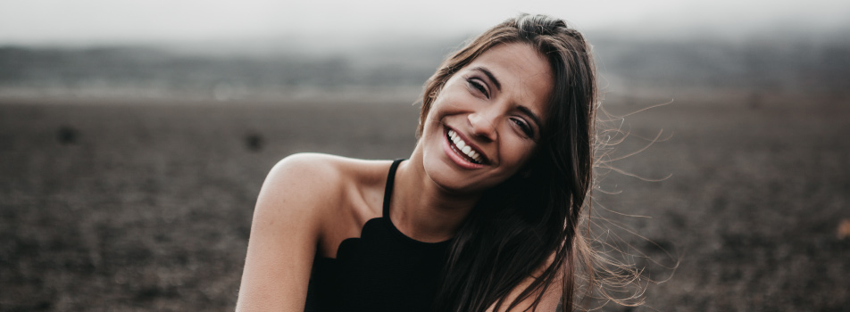 Brunette woman. wearing a black tank top smiles while leaning her head to one side and sitting in a dirt field