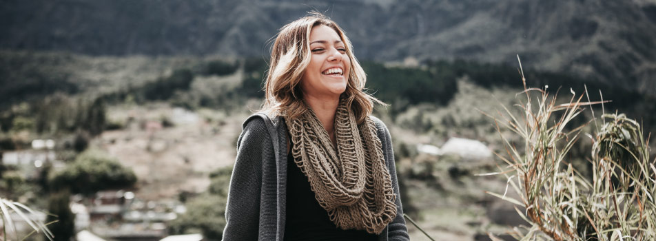 Brunette woman wearing a beige scarf smiles with healthy gums as she stands at the foot of a mountain