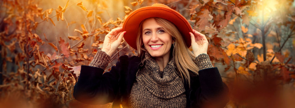 Blonde middle-aged woman wearing a black sweater and rust-colored hat smiles with dentures in the fall leaves