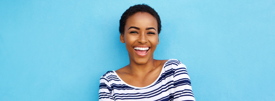 Dark-haired woman with a navy and white striped blouse smiles while standing against a sky blue wall