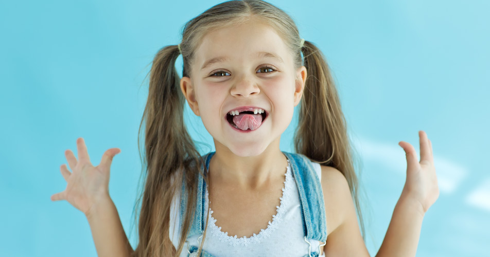 Brunette little girl wearing overalls smiles showing off her missing front teeth