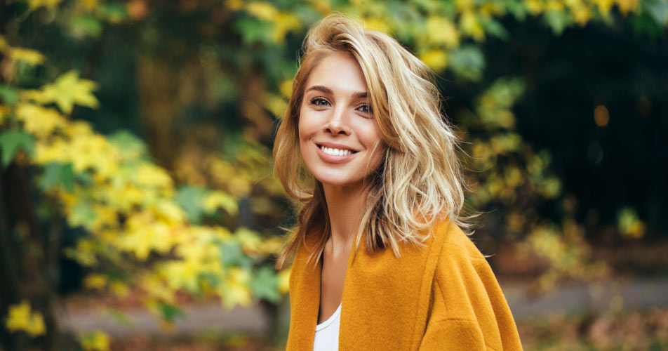Blonde woman with dental implants smiles outside while wearing a yellow cardigan