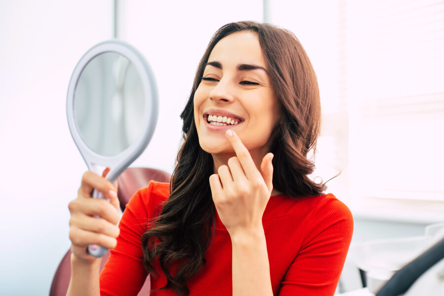 Brunette woman in a red shirt smiles in a mirror with her new dentures