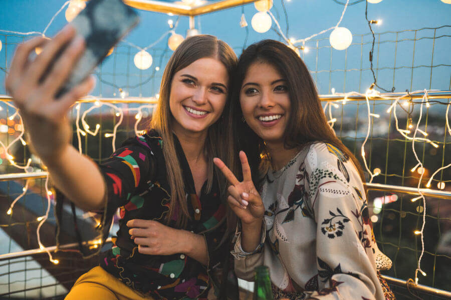 A blonde and a brunette woman smile like celebrities while taking a selfie in front of twinkle lights