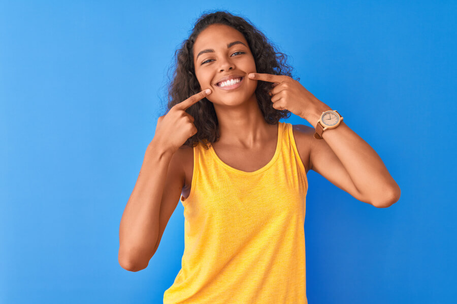 Brunette woman in a yellow tanktop points at her healthy smile against a blue wall