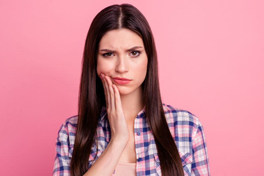 Brunette woman in a plaid shirt cringes in pain against a pink background due to a dental abscess
