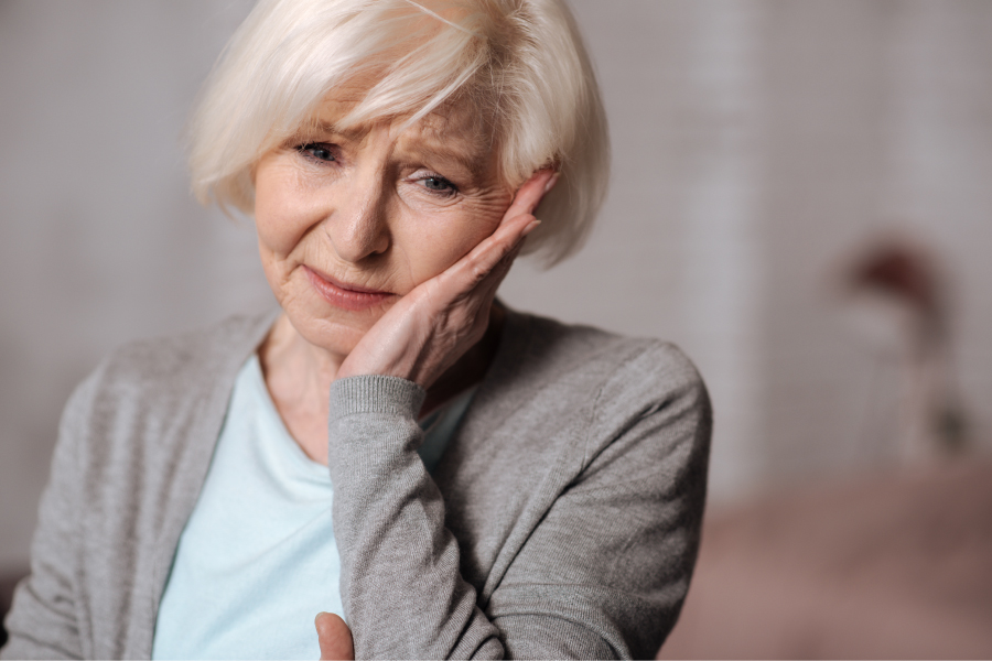 White-haired woman cringes in pain and touches her cheek because she needs root canal therapy