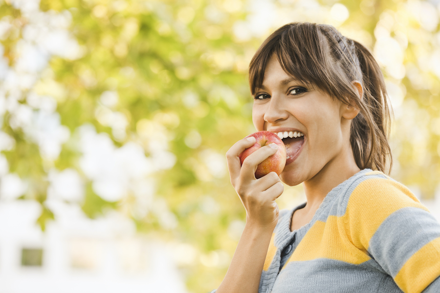 Smiling brunette white woman in a yellow and gray striped shirt eats an apple outside