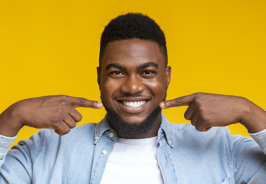 Black man smiles while pointing at his teeth against a yellow background